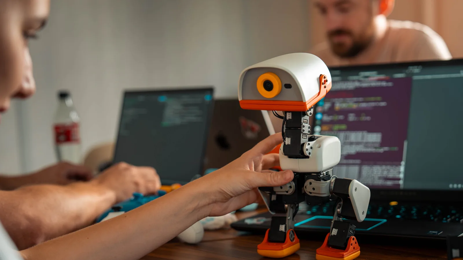 A hand holding Microduck upright on a desk between two laptops
